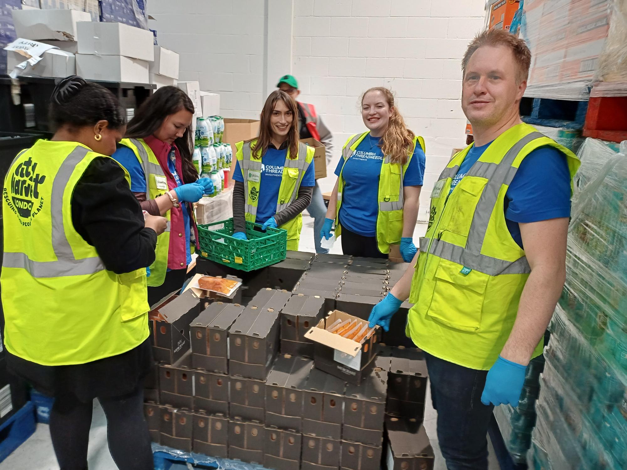 People wearing safety vests and gloves stand around stacked boxes on pallets, sorting packaged food items inside a warehouse space